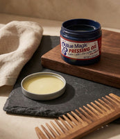 Blue Magic Pressing Oil - barattolo aperto su ardesia con pettine di legno, l'olio giallo visibile